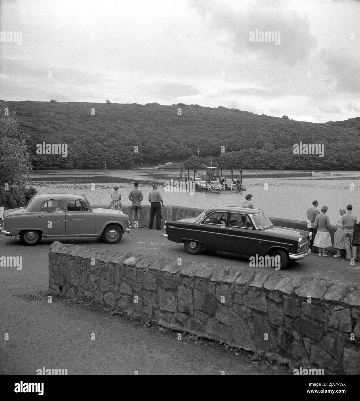 1950s, storico, persone e automobili dell'epoca, tra cui un Austin Cambridge, in attesa a Feock, guardando il King Harry Ferry, attraversando le strade Carrick raggiungere l'estuario del fiume Fal in Cornovaglia, Inghilterra, Regno Unito. Un piccolo ponte di traghetti a catena è stato istituito su questa parte dell'estuario nel 1888 utilizzando un motore a vapore per tirarlo lungo le catene e questo è stato utilizzato fino all'introduzione di un motore a bisello nel 1956. Uno dei viaggi in traghetto più panoramici al mondo, è ora uno dei soli cinque traghetti della catena in Inghilterra. Foto Stock