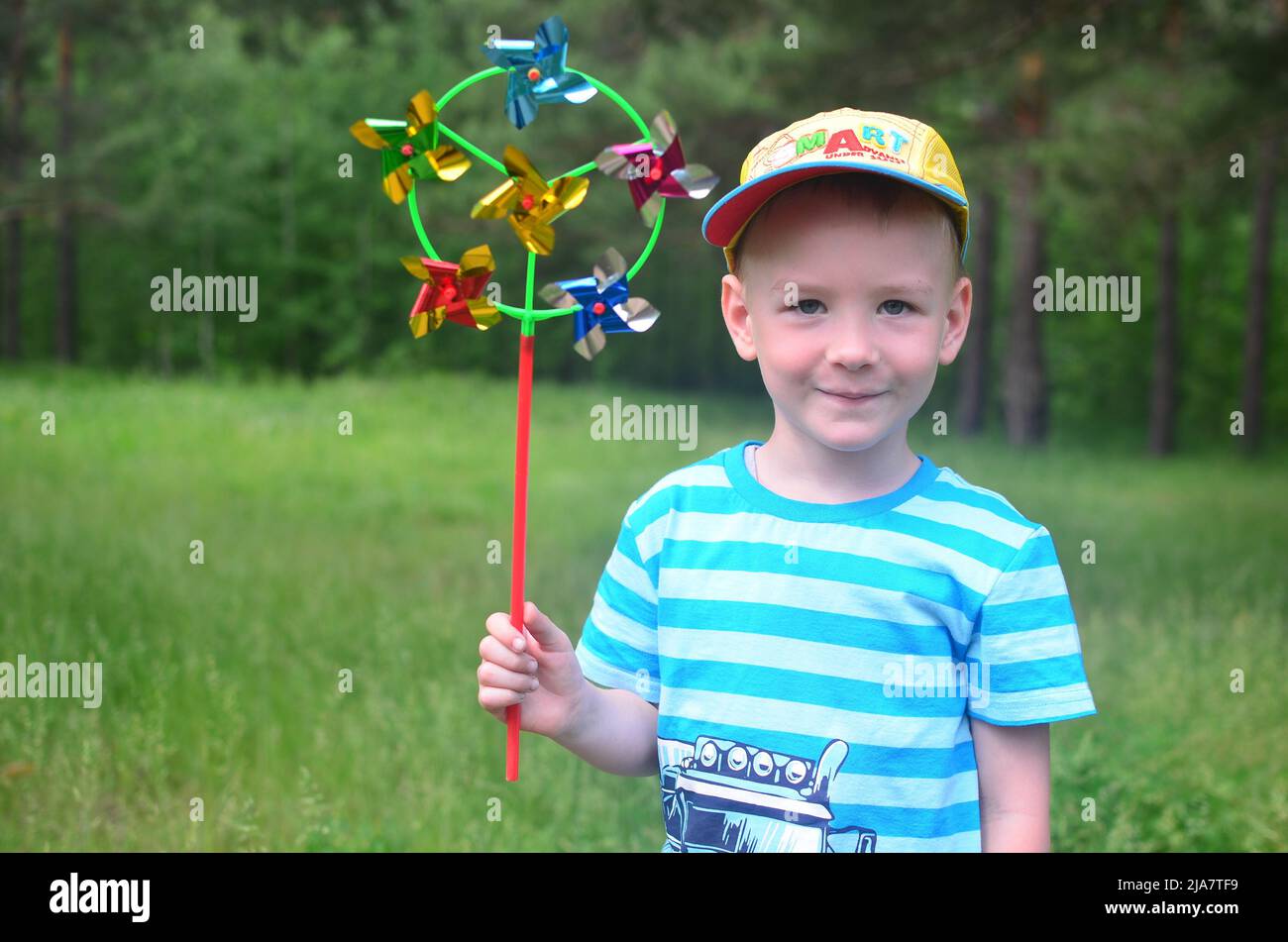 felice infanzia - concetto. Il ragazzo caucasico sorride in un cappellino e una t-shirt a righe con un mulino a vento in mano sullo sfondo di una foresta verde. Foto Stock