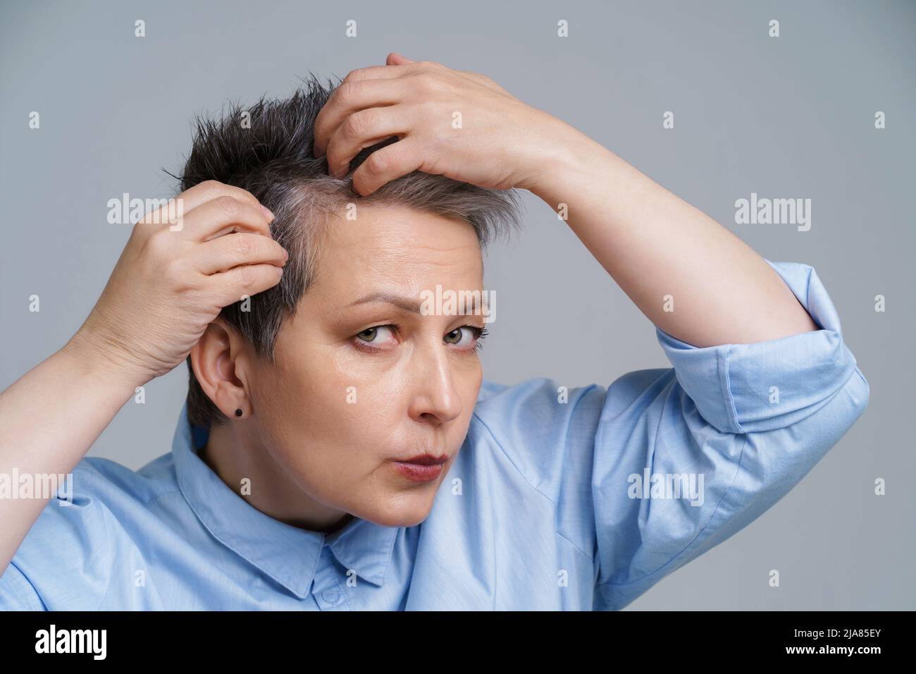 Ritratto di donna matura nel 50s controllando i suoi capelli e infelice vedere un risultato a specchio. Bella donna grigia con capelli che si occupa di dandruff problema di controllo nello specchio. Foto Stock