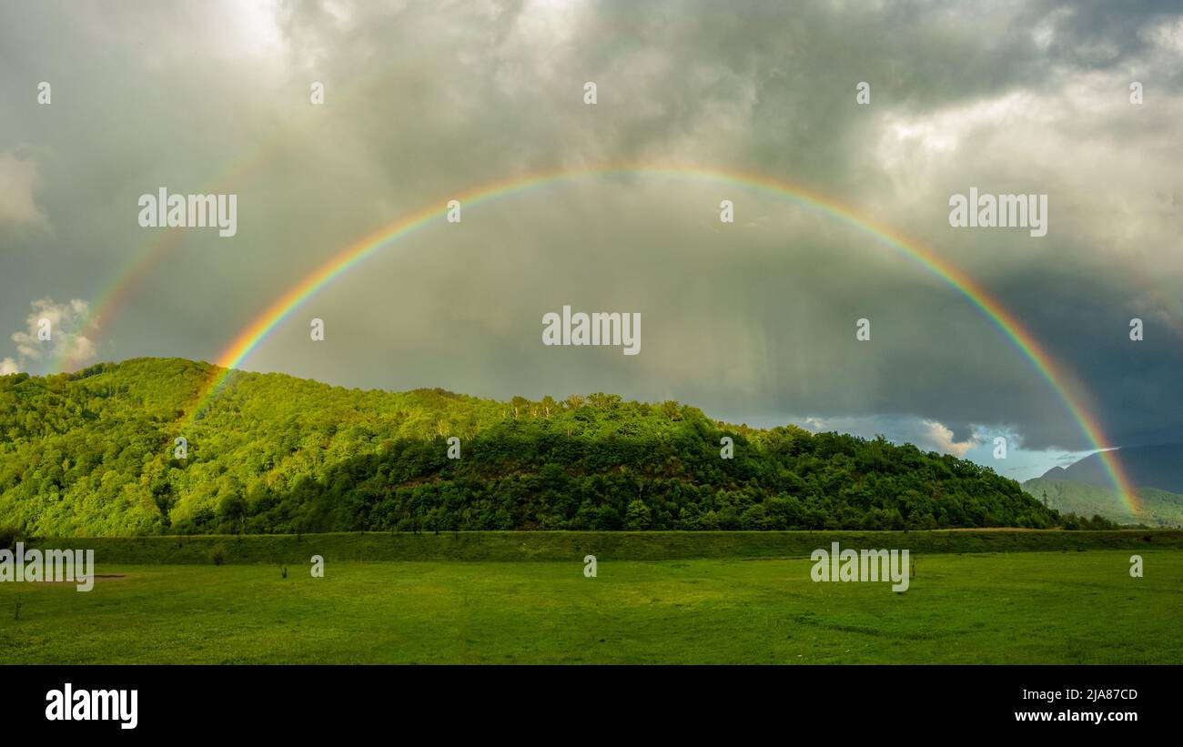 Un arcobaleno colorato e ad arco che appare sopra un'area rurale durante una giornata di pioggia. La foresta è illuminata dal fenomeno colorato e dalla luce del sole. Foto Stock