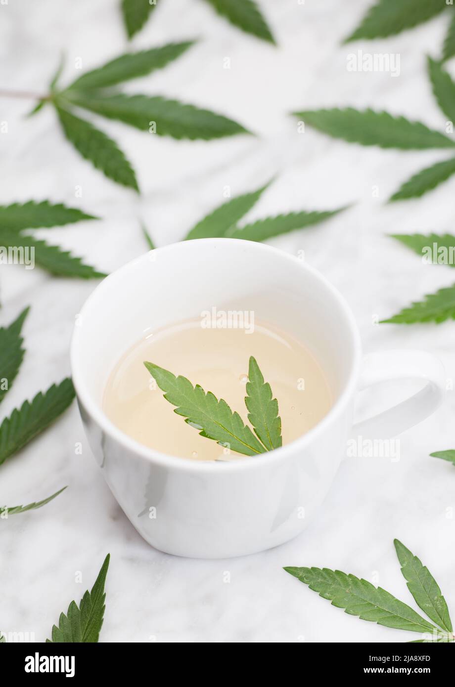 tazza bianca con infusione di marijuana con foglia verde all'interno su tavolo di marmo con foglie. vista dall'alto. Foto Stock