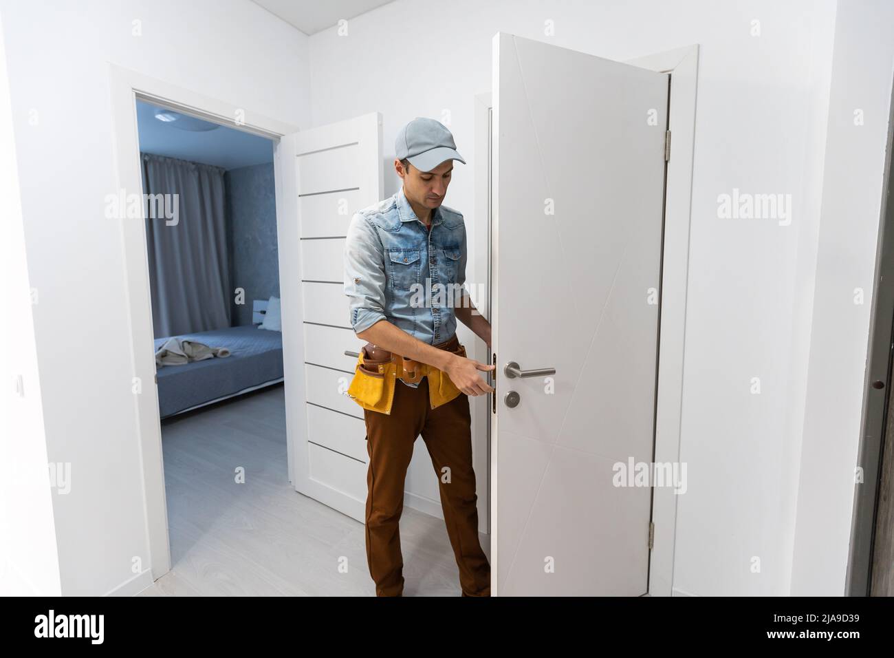 Montaggio di una serratura sulla porta di ingresso in legno anteriore. Ritratto di giovane fabbro operaio in uniforme blu montaggio pomello porta. Professionale Foto Stock