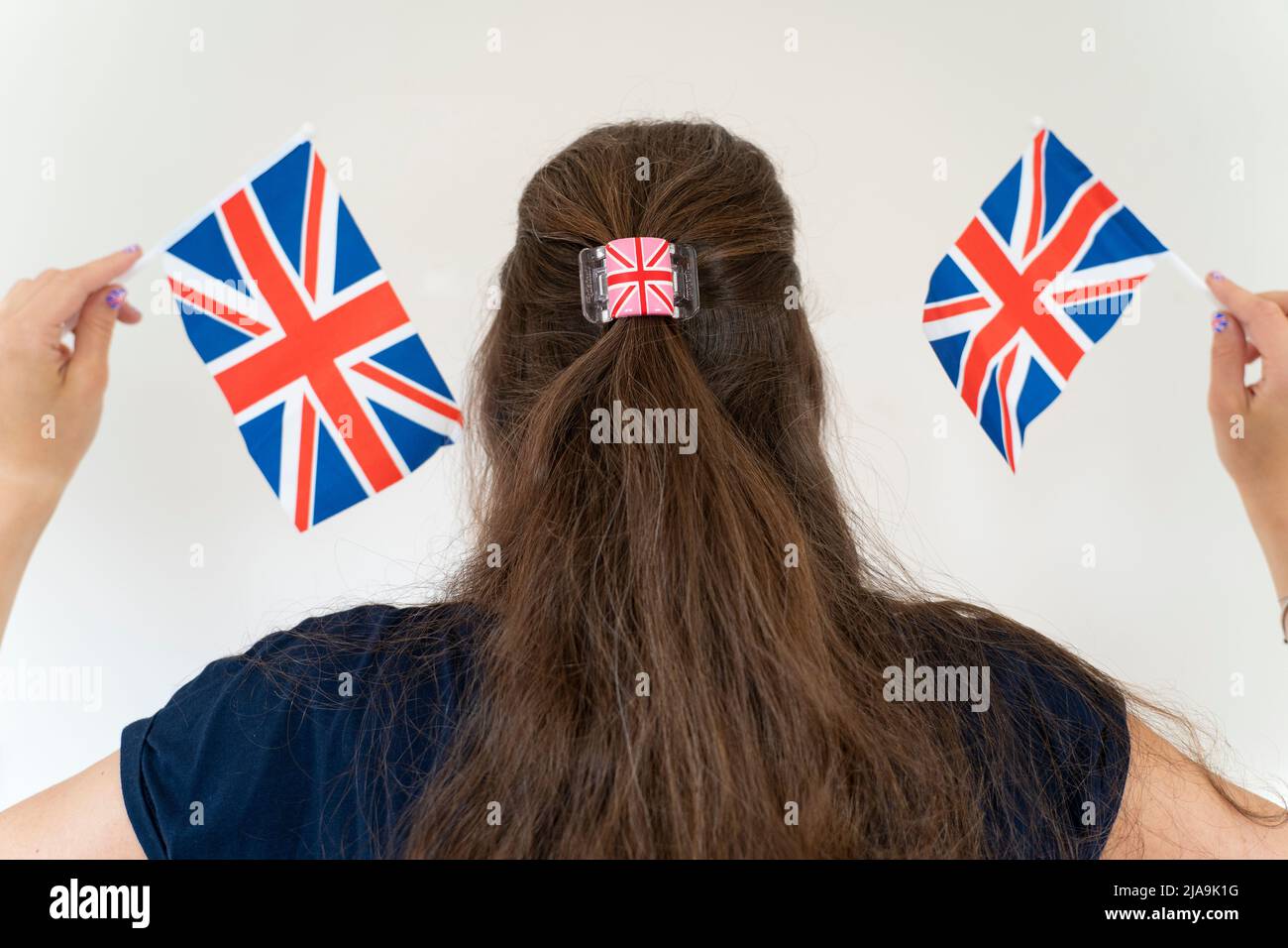 Fan femmina che tiene e agita le bandiere britanniche e indossa una clip per capelli con Union Jack. Tema - Giubileo del platino Elisabetta II giugno 2022 Foto Stock