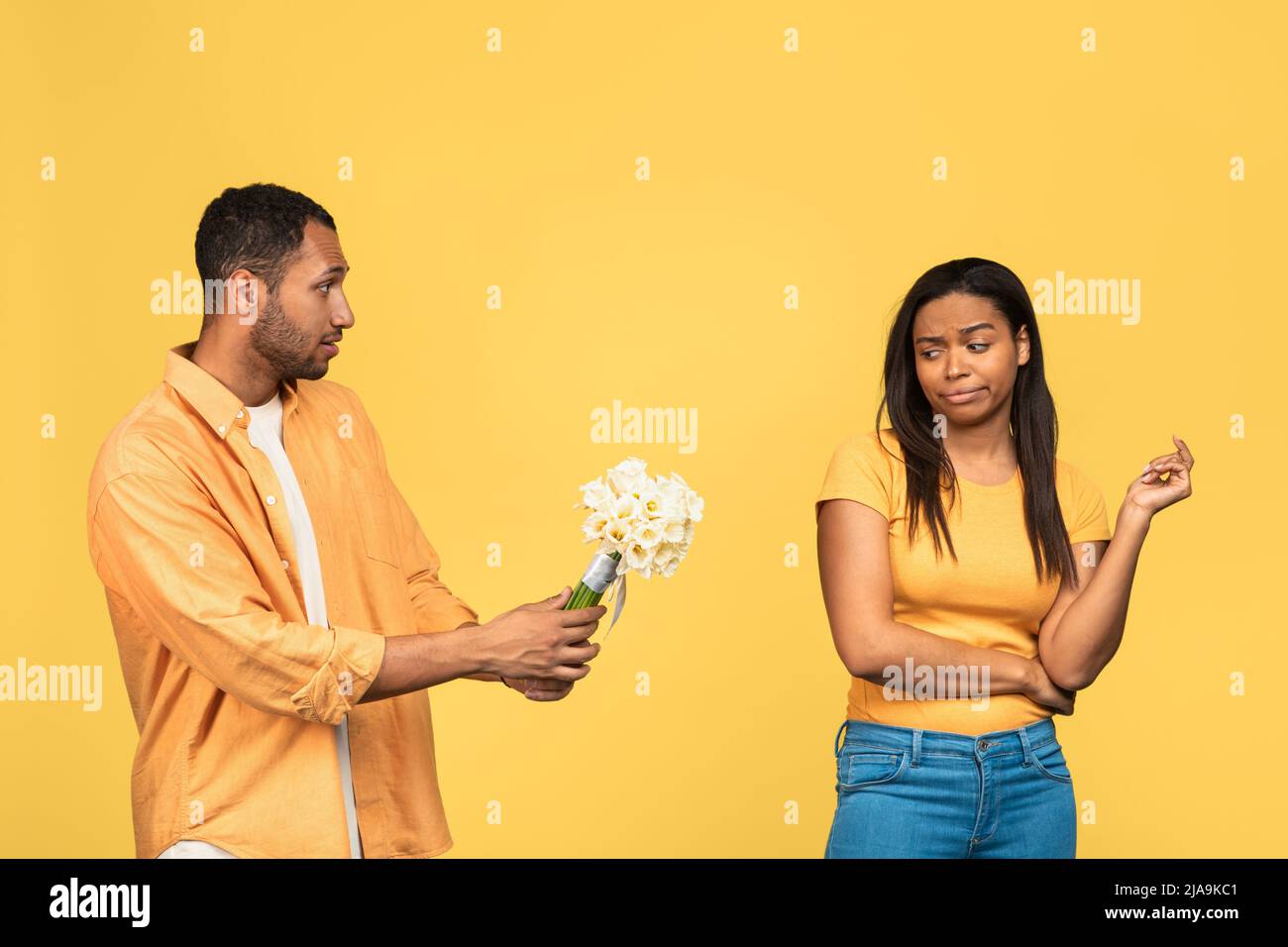 Ragazza nera arrabbiata rigettando i fiori e le scuse del suo ragazzo triste dopo la lotta su sfondo giallo studio Foto Stock