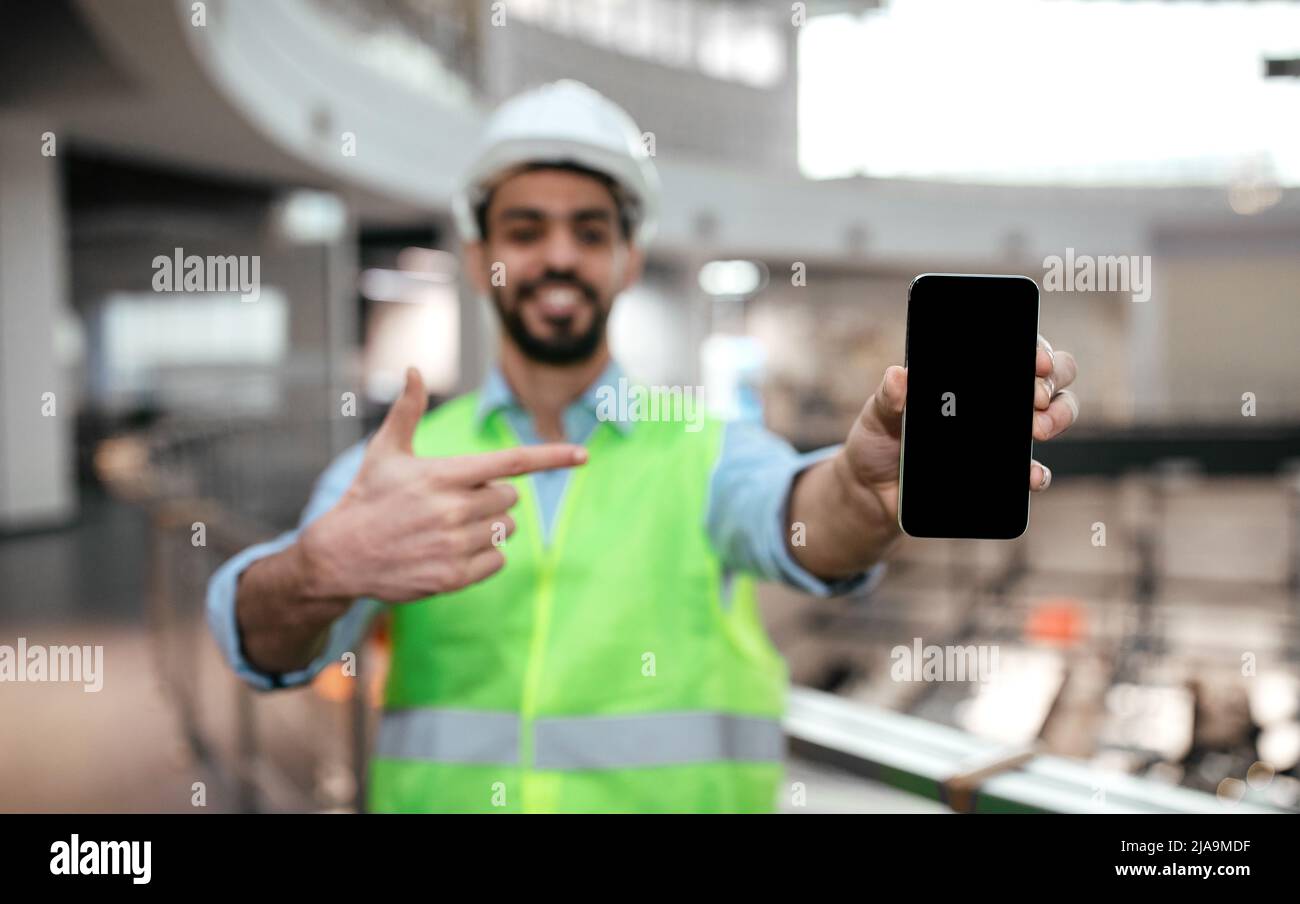 Felice ingegnere islamico millenario maschio in uniforme protettiva, elmetto con punta di barba dito al telefono Foto Stock