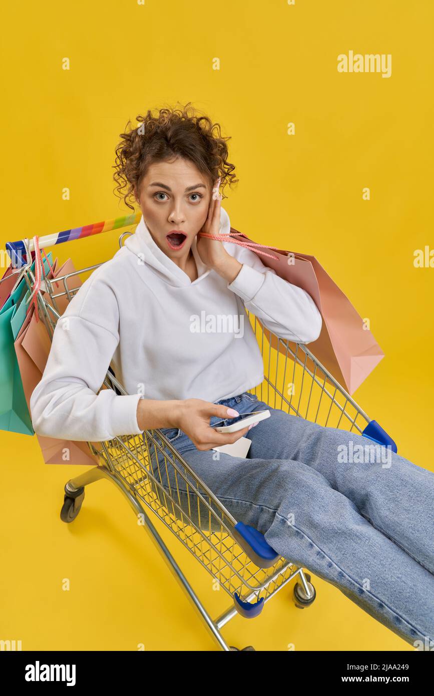 Confuso ragazza che tiene il telefono, controllando i costi, mentre seduto nel carrello della spesa. Sopra la vista della donna sorpresa che tiene la mano a faccia nel carrello del negozio, isolato su sfondo arancione. Concetto di spesa. Foto Stock
