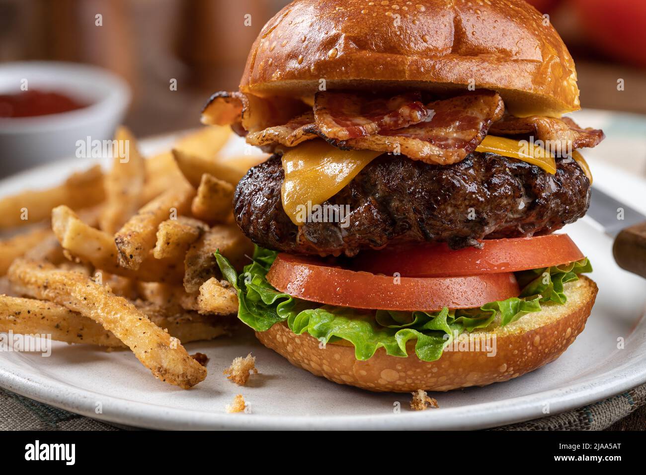 Primo piano di un cheeseburger con pancetta e lattuga e pomodoro su una panna tostata e patatine fritte su un piatto Foto Stock