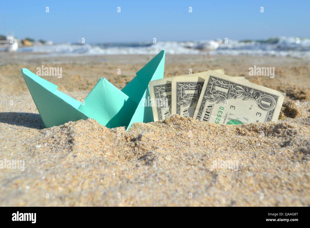 Piccola barca di carta blu e carta da tre dollari bollette mezzo in sabbia sulla spiaggia vicino al mare in estate giorno di sole. Concetto di viaggio, sogni, spese, denaro, finanza, turismo, riposo, vacanza obiettivi desidera Foto Stock