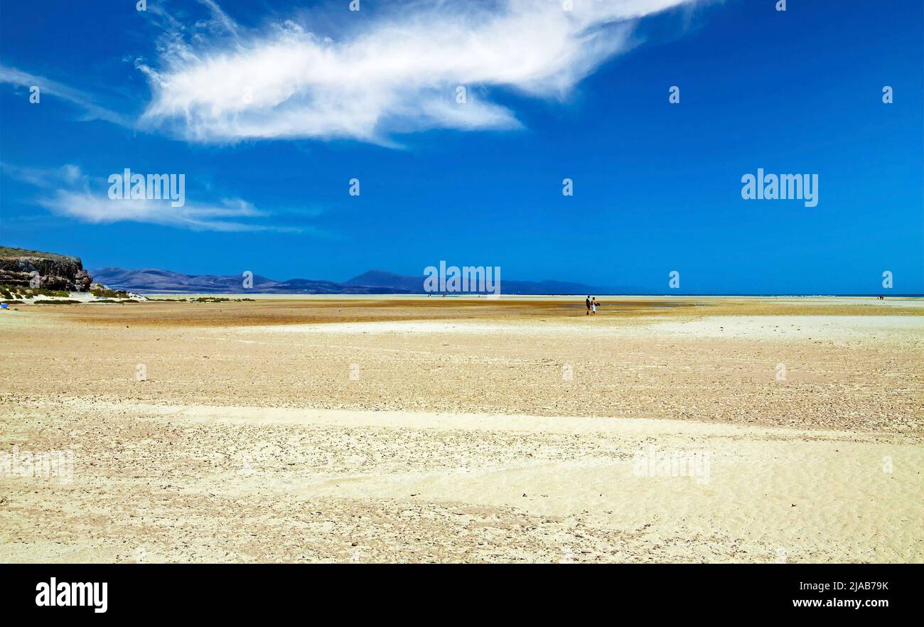 Ampia area di sabbia infinita durante la bassa marea ebb, silhouette di poche persone, piccola striscia blu ocena, colline, cielo - Sotavento Beach, Fuerteventura Foto Stock