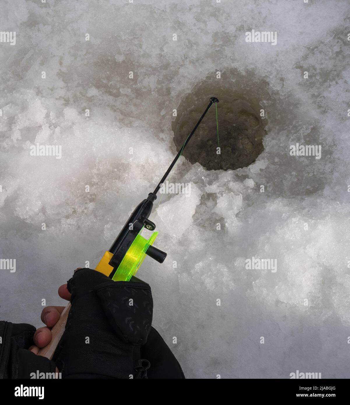 Mani con guanti che tengono una canna da pesca sopra un foro nel ghiaccio che copre un fiume congelato. Foto Stock