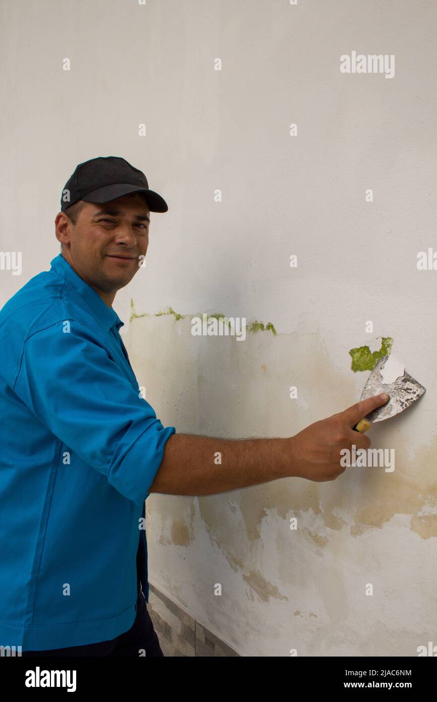 Immagine di un uomo sorridente che rimuove la vernice da una parete con una spatola per ripristinarla dalla muffa. Foto Stock