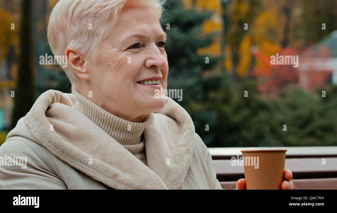 Felice sognante grigio-capelli anziani matura donna nonna seduta sul banco del parco bere caffè caldo tè da tazza usa e getta sano cheerful senior Foto Stock
