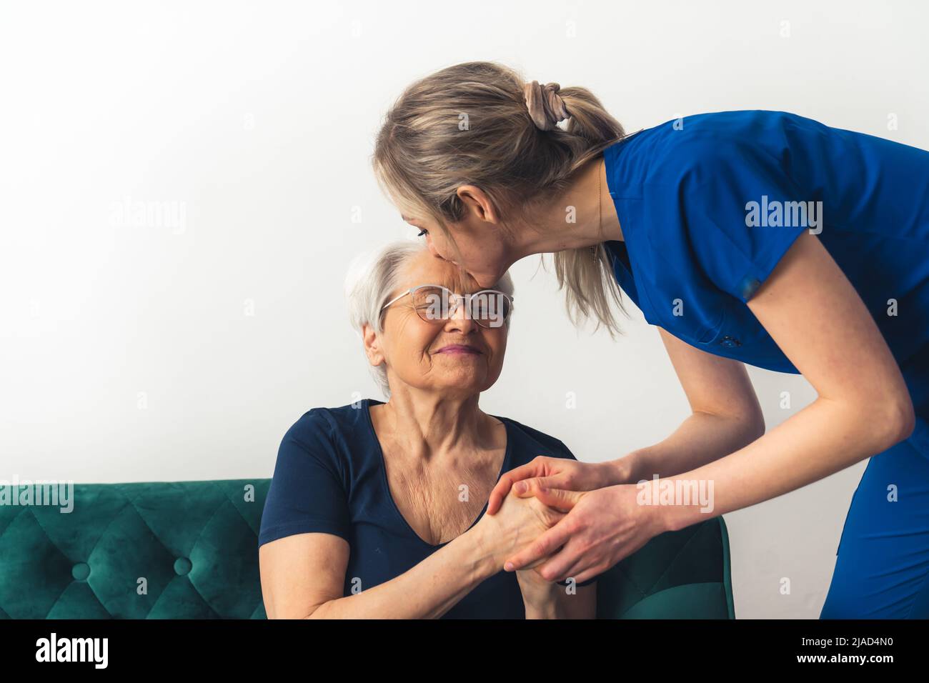 Donna MD nel suo 20s indossare scrub blu appoggiata verso una donna anziana seduta su un divano, baciandola sulla fronte. Scatto al coperto. Foto di alta qualità Foto Stock