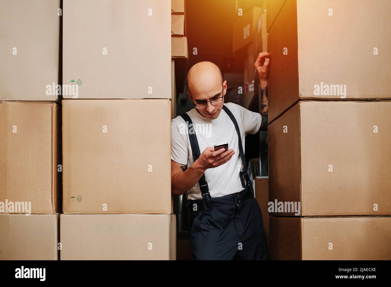 Uomo coraggioso e stanco addetto al magazzino che si appoggia su una pila di scatole, guardando il suo telefono. È in piedi fra le pile alte delle scatole di cartone. Foto Stock