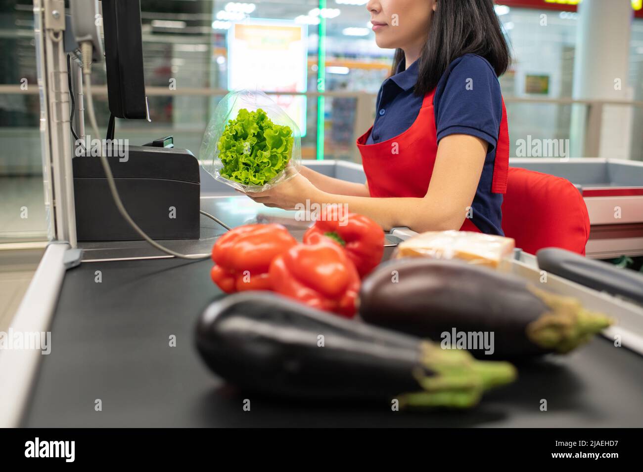 Donna asiatica che indossa grembiule rosso seduto alla cassa che emette un segnale acustico verdure Foto Stock