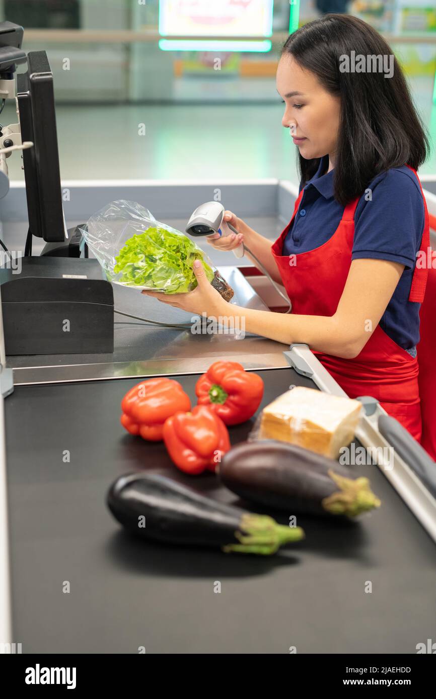 Immagine verticale di attraente cassiere asiatico che indossa grembiule rosso vendendo verdure fresche al cliente Foto Stock