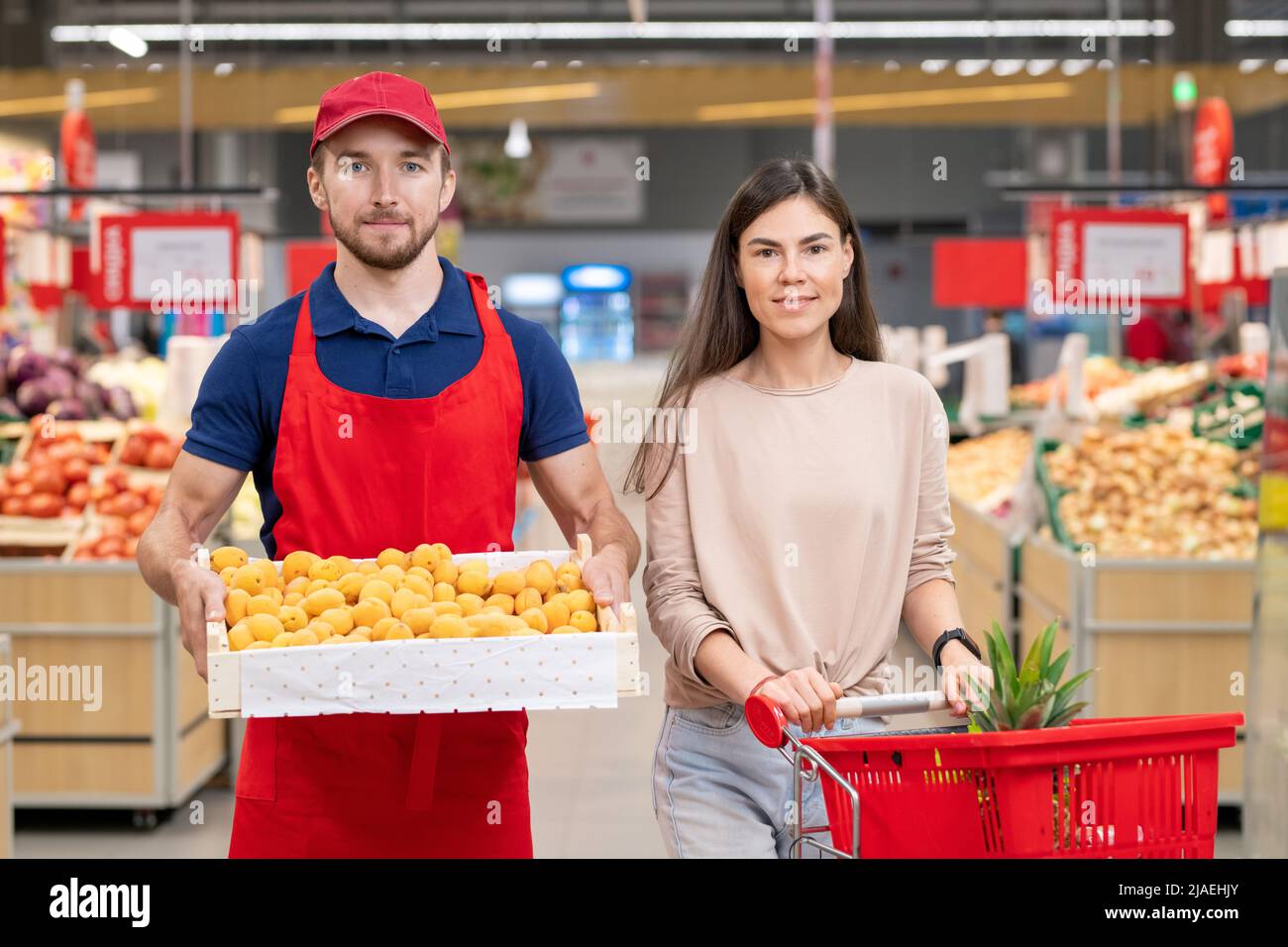 Colpo medio orizzontale di lavoratore maschio che tiene scatola di albicocche stare in piedi con una giovane cliente femminile nella corsia del supermercato Foto Stock