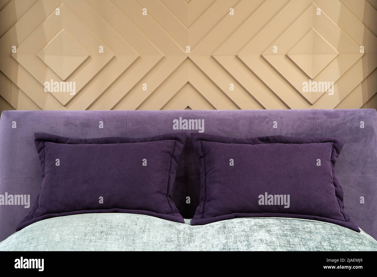 Vista ravvicinata di cuscini viola e testiera di un letto matrimoniale vuoto con copriletto grigio. Parete di design con stampaggio volumetrico in stucco beige in fo Foto Stock