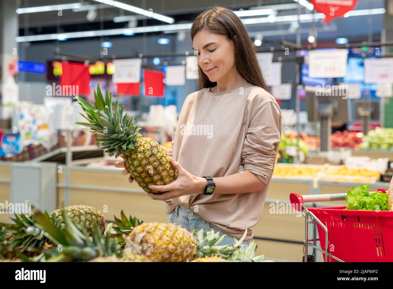 Giovane donna che indossa abiti casual scegliendo il migliore ananas nel moderno supermercato, spazio di copia Foto Stock