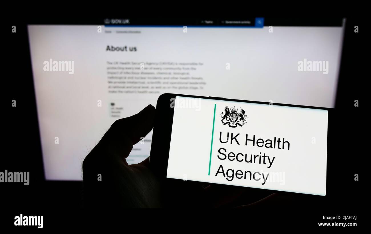 Persona che detiene il telefono cellulare con il logo dell'agenzia UK Health Security Agency (UKHSA) sullo schermo di fronte alla pagina web. Mettere a fuoco sul display del telefono. Foto Stock