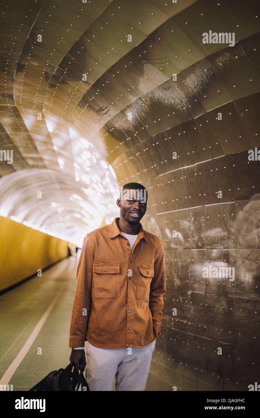 Una borsa da trasporto sorridente da uomo mentre si alza con la mano in tasca al tunnel della metropolitana Foto Stock