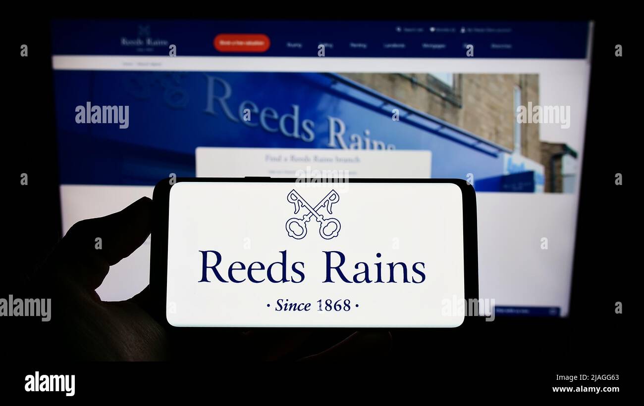Persona che tiene il cellulare con il logo della società immobiliare britannica Reeds Rains Limited sullo schermo di fronte al sito web. Mettere a fuoco sul display del telefono. Foto Stock