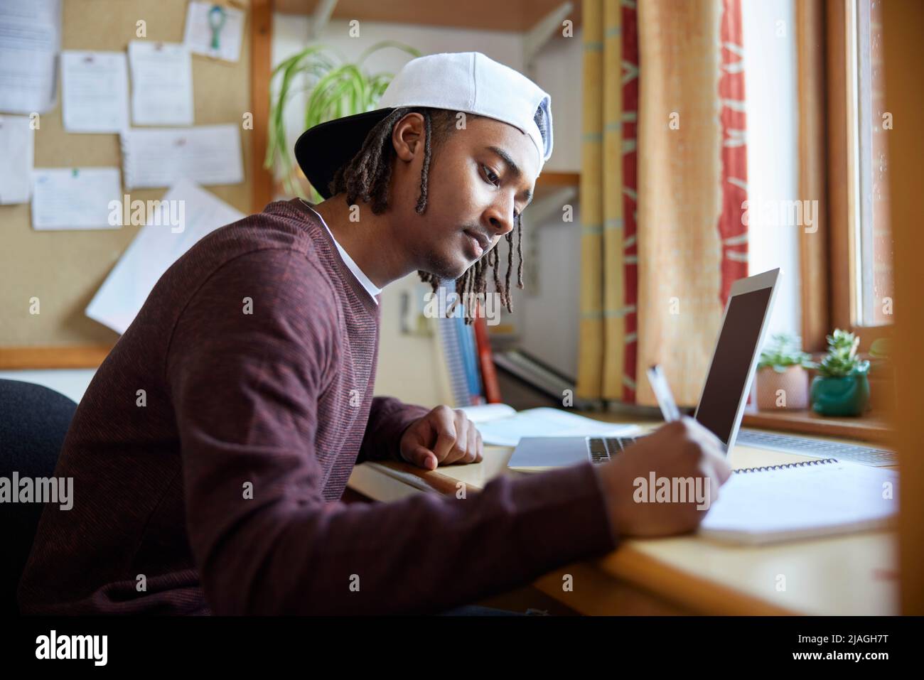 Studente universitario maschile o universitario che indossa il cappellino da baseball che studia con il computer portatile alla scrivania in camera Foto Stock