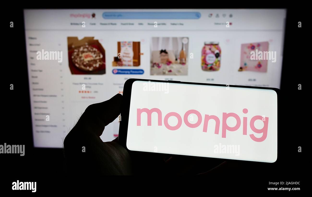 Persona che tiene smartphone con il logo della società di biglietti d'auguri Moonpig Group plc sullo schermo di fronte al sito web. Mettere a fuoco sul display del telefono. Foto Stock