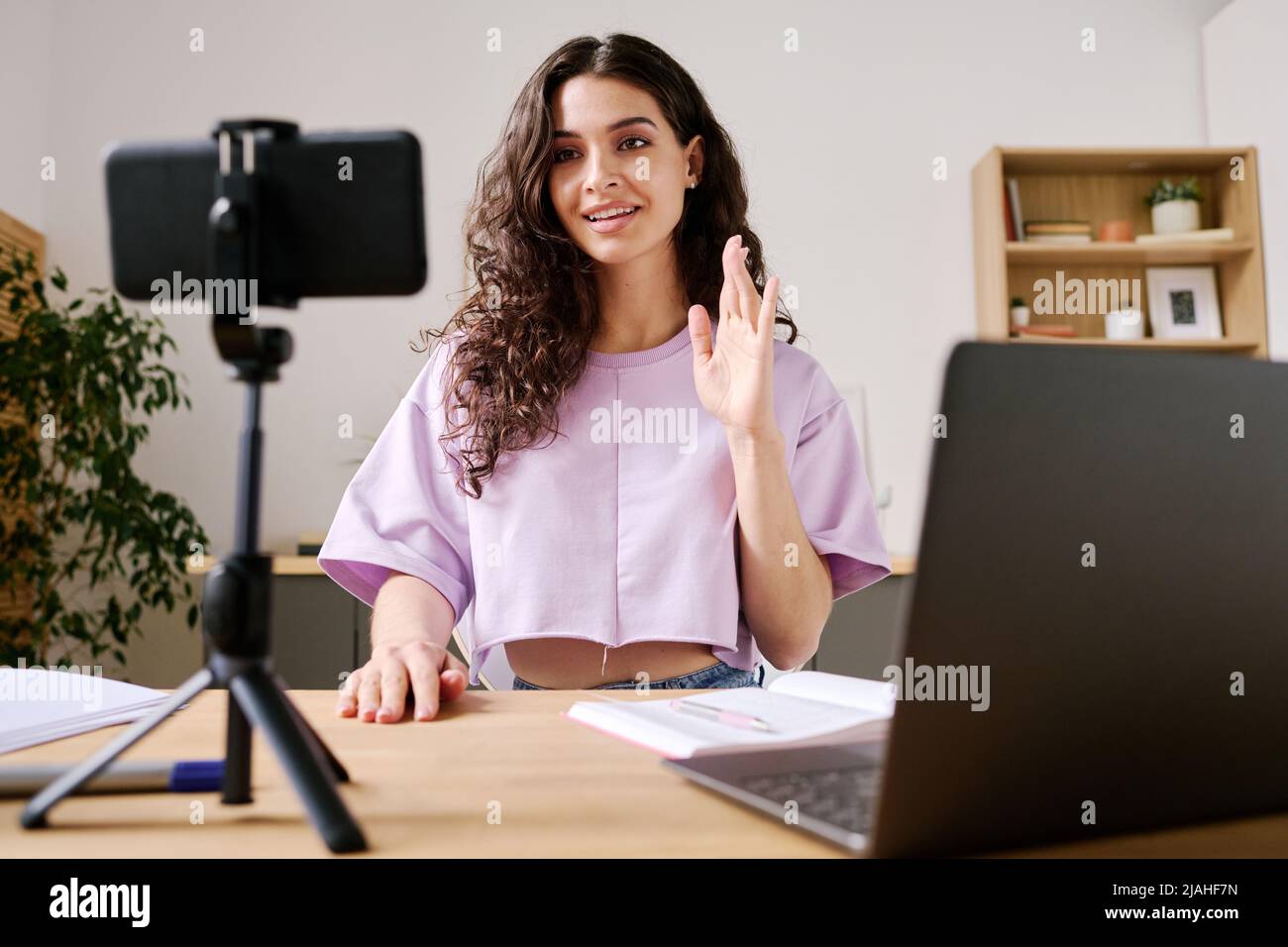 Giovane blogger femminile seduto alla scrivania di fronte allo smartphone su treppiede video di partenza con il saluto dei suoi seguaci Foto Stock