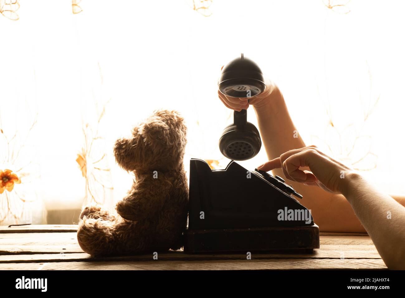 La mano di una donna compone un numero di telefono su un vecchio telefono e un orsacchiotto si siede accanto ad esso a casa sul tavolo, un telefono retrò è nero, una chiamata Foto Stock