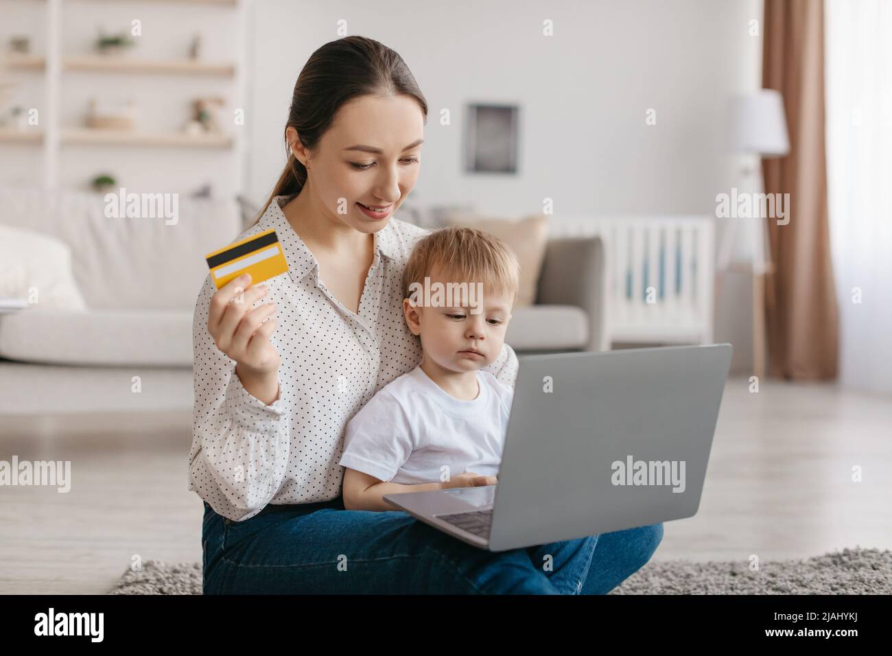 Felice madre giovane e suo figlio del bambino che acquista in linea con il laptop e la carta di credito, ragazzo del capretto che si siede sui giri della mamma Foto Stock