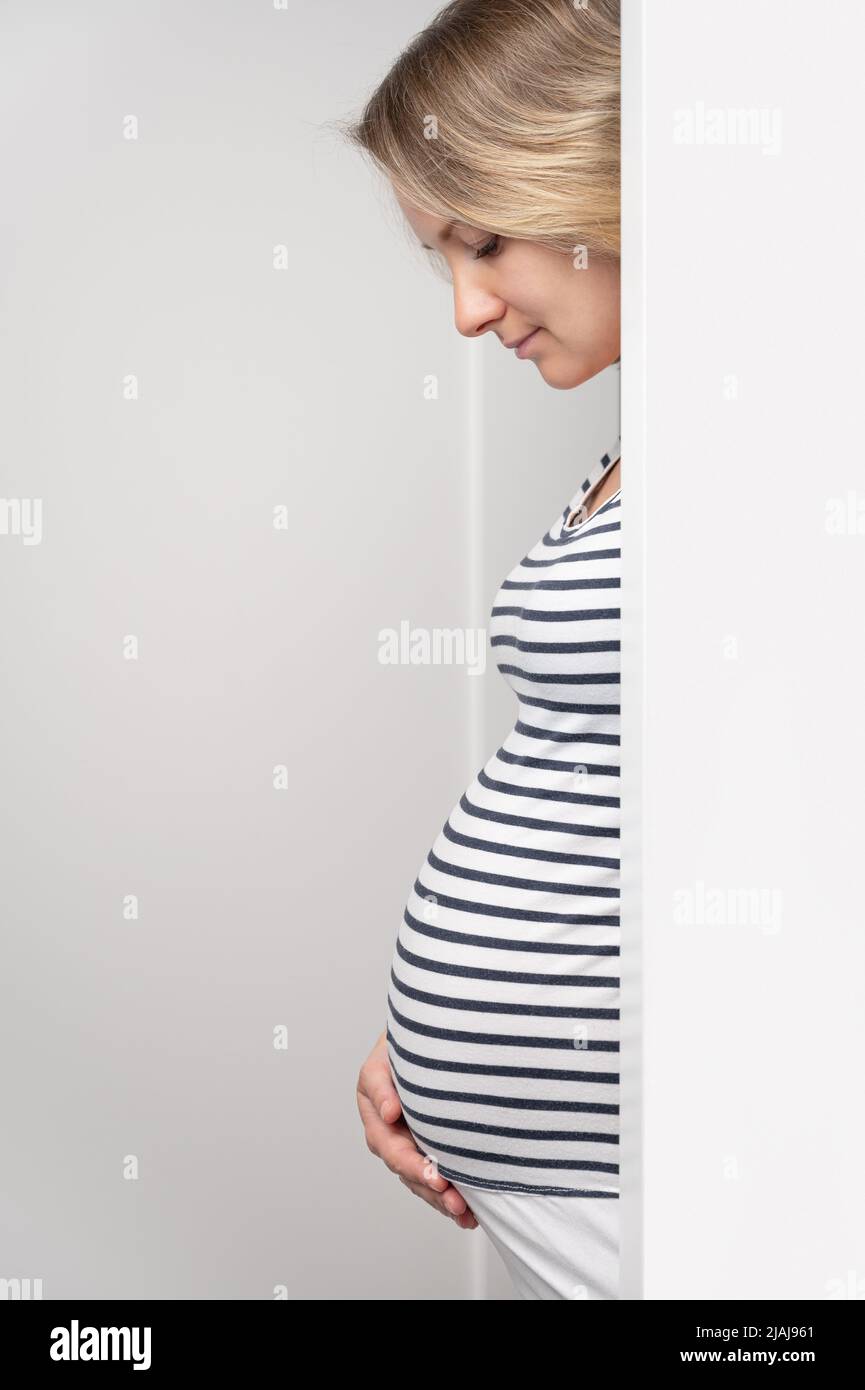 Donna incinta che sbucciava dalla parete bianca. Donna incinta felice che tocca teneramente il suo ventre. Aspettativa del bambino. Foto Stock