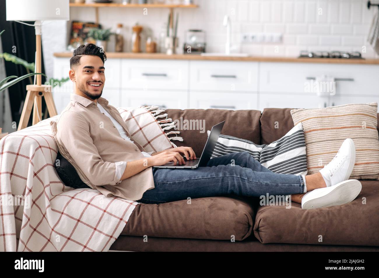 Attraente uomo arabo sorridente o indiano in abbigliamento casual elegante, freelancer o studente, seduto sul divano in salotto, lavorare su un progetto utilizza un laptop, guardando la macchina fotografica, sorridendo felicemente Foto Stock