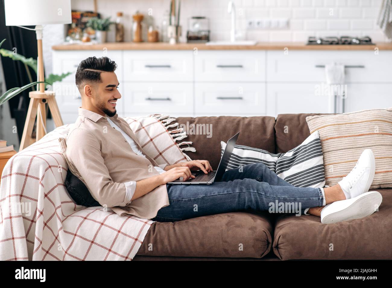 Bel ragazzo alla moda arabo o indiano in abiti casual elegante, freelancer o studente, seduto sul divano in un soggiorno, utilizza un computer portatile per lavorare su un progetto, elearning, navigare in internet, sorridendo Foto Stock