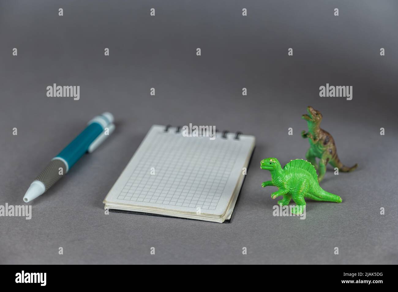 Due miniature Dinosaur, notebook e penna a sfera su sfondo grigio. Piccole figure verdi di animali di preda in piedi sulle loro zampe posteriori. SPR Foto Stock