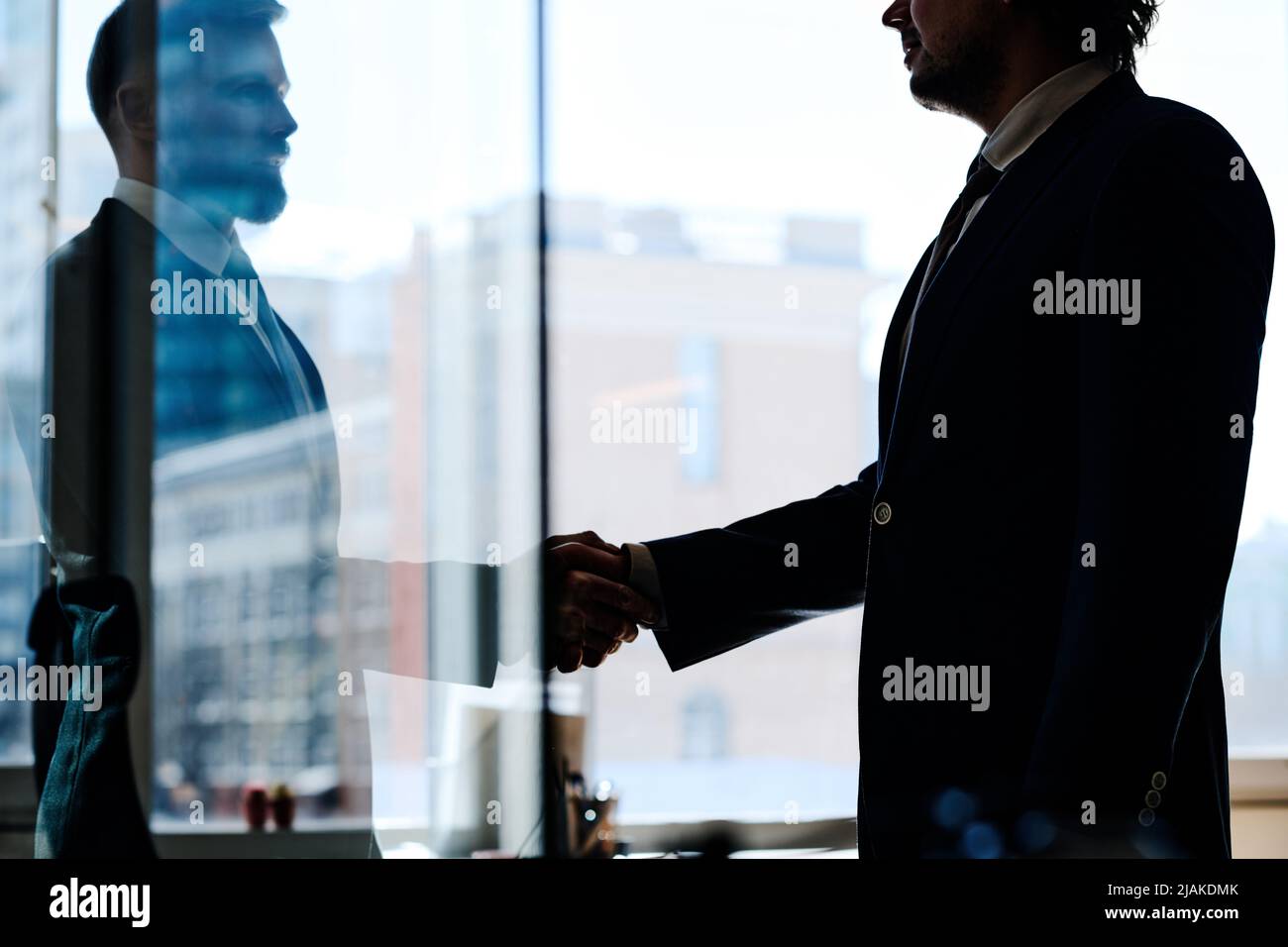 Due uomini d'affari in tute scuotendo le mani per concludere un affare in ufficio in piedi dietro il muro di vetro Foto Stock