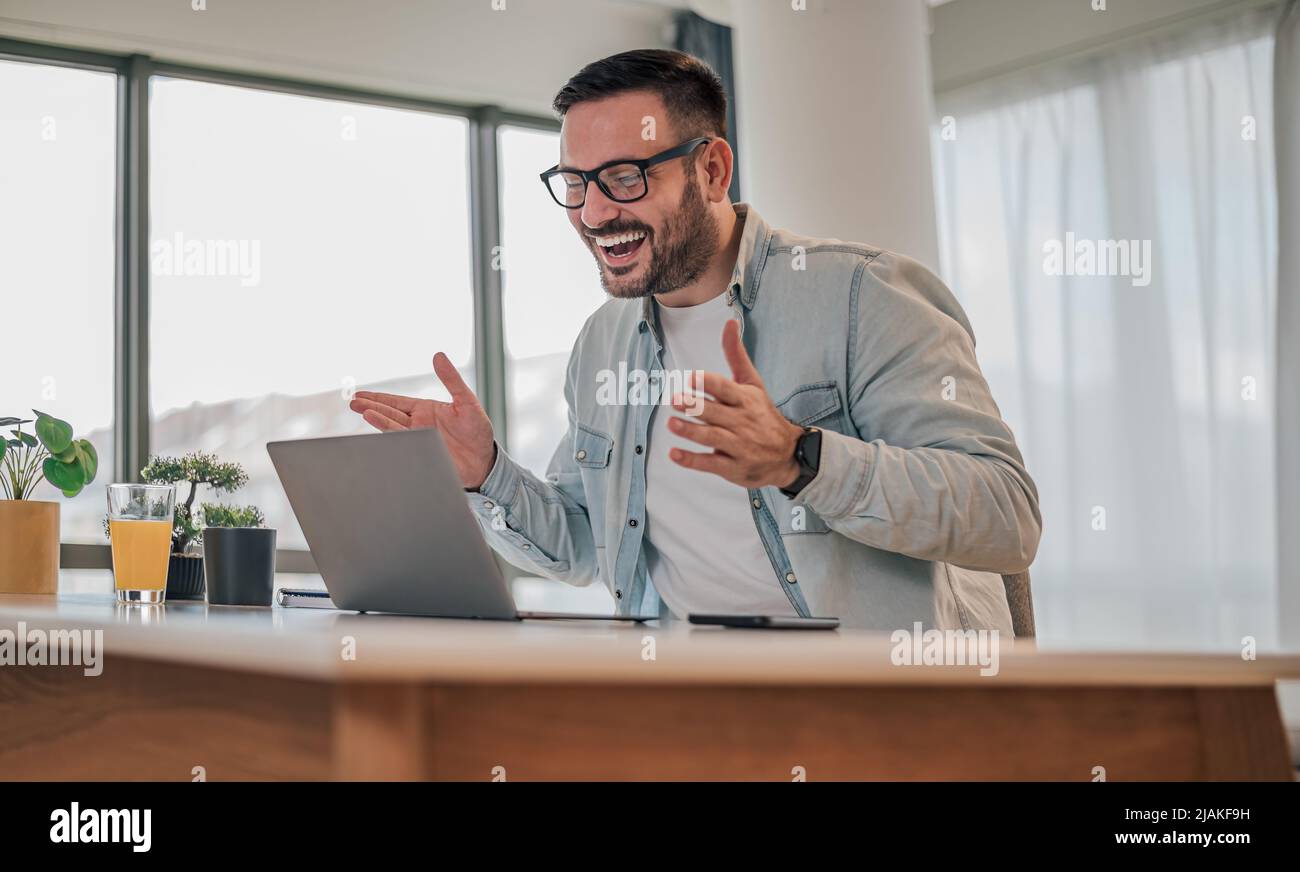 Un uomo d'affari felice che gesturing mentre guarda il laptop. Giovane professionista maschile indossa casuals intelligenti. Lavora alla scrivania in ufficio. Foto Stock