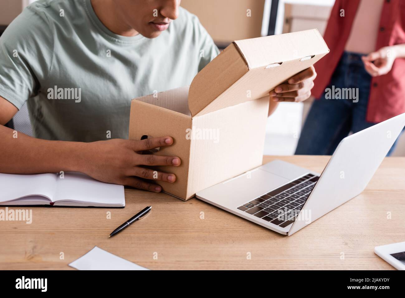 Vista ritagliata del venditore afro-americano che tiene la scatola di cartone vicino al laptop nel negozio in linea di fotoricettore Foto Stock