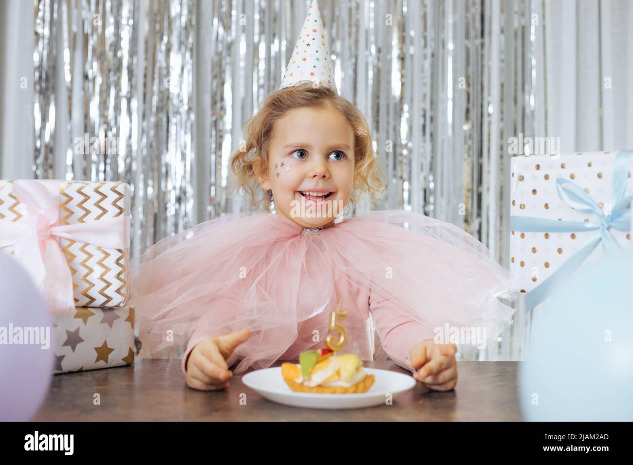 Momenti felici. Emozioni positive dei bambini. Sorridente ragazza con occhi luminosi in vestito festivo è seduta di fronte alla torta di compleanno decorata con Foto Stock