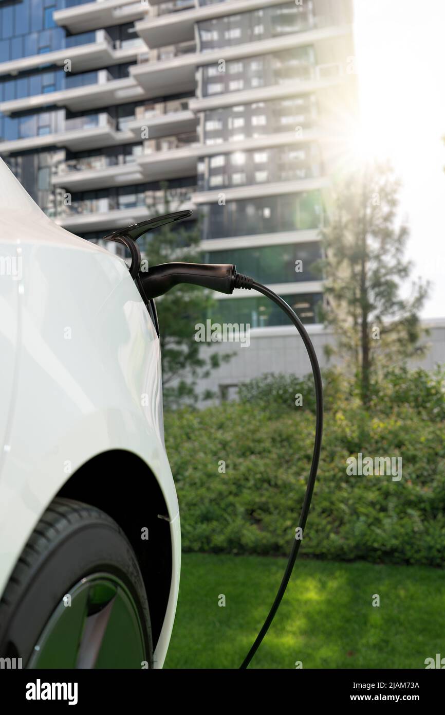 Primo piano dell'auto elettrica con un cavo di ricarica collegato sullo sfondo di una città ecologica verde Foto Stock