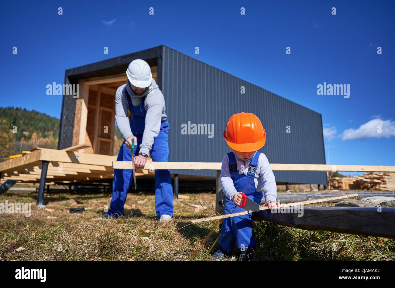 Padre con bambino figlio costruzione in legno cornice casa in stile scandinavo. Costruttori maschi che utilizzano la sega a mano per tagliare tavole in cantiere il giorno di sole. Carpenteria e concetto di famiglia. Foto Stock