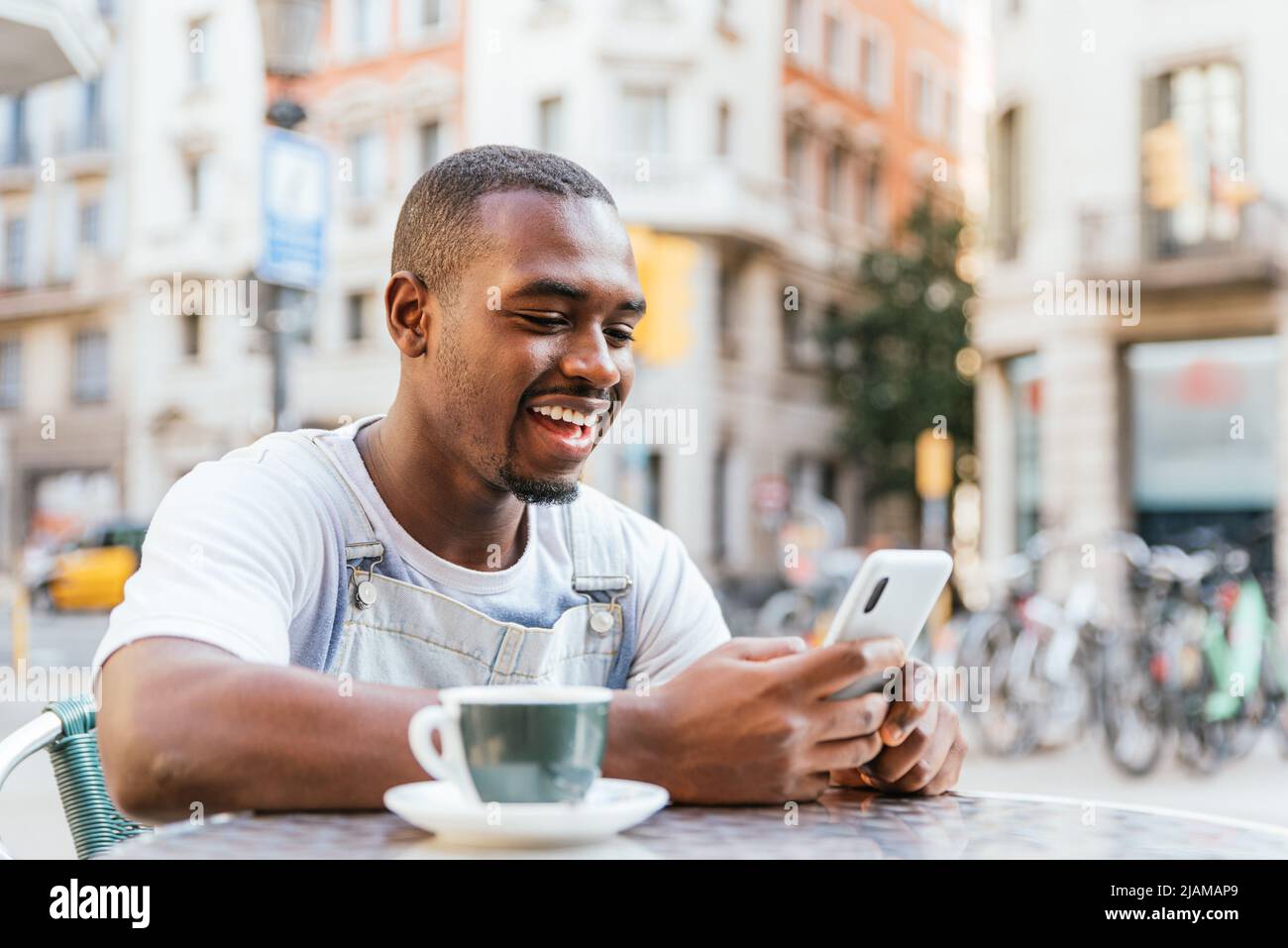 Allegro giovane africano americano maschile millenario in abiti casual. Sorride felicemente mentre legge le buone notizie sullo smartphone seduto al tavolo con una tazza di caffè in Street cafe Foto Stock