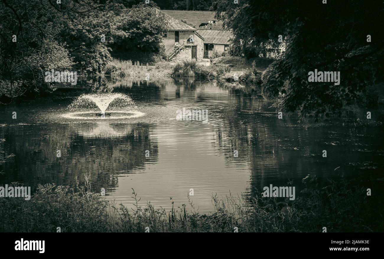 Vista sull'acqua ferma con fontana da gioco. Vecchia fattoria sullo sfondo riflessa nel lago circondato dal verde. Tranquillo, senza tempo, bello Foto Stock