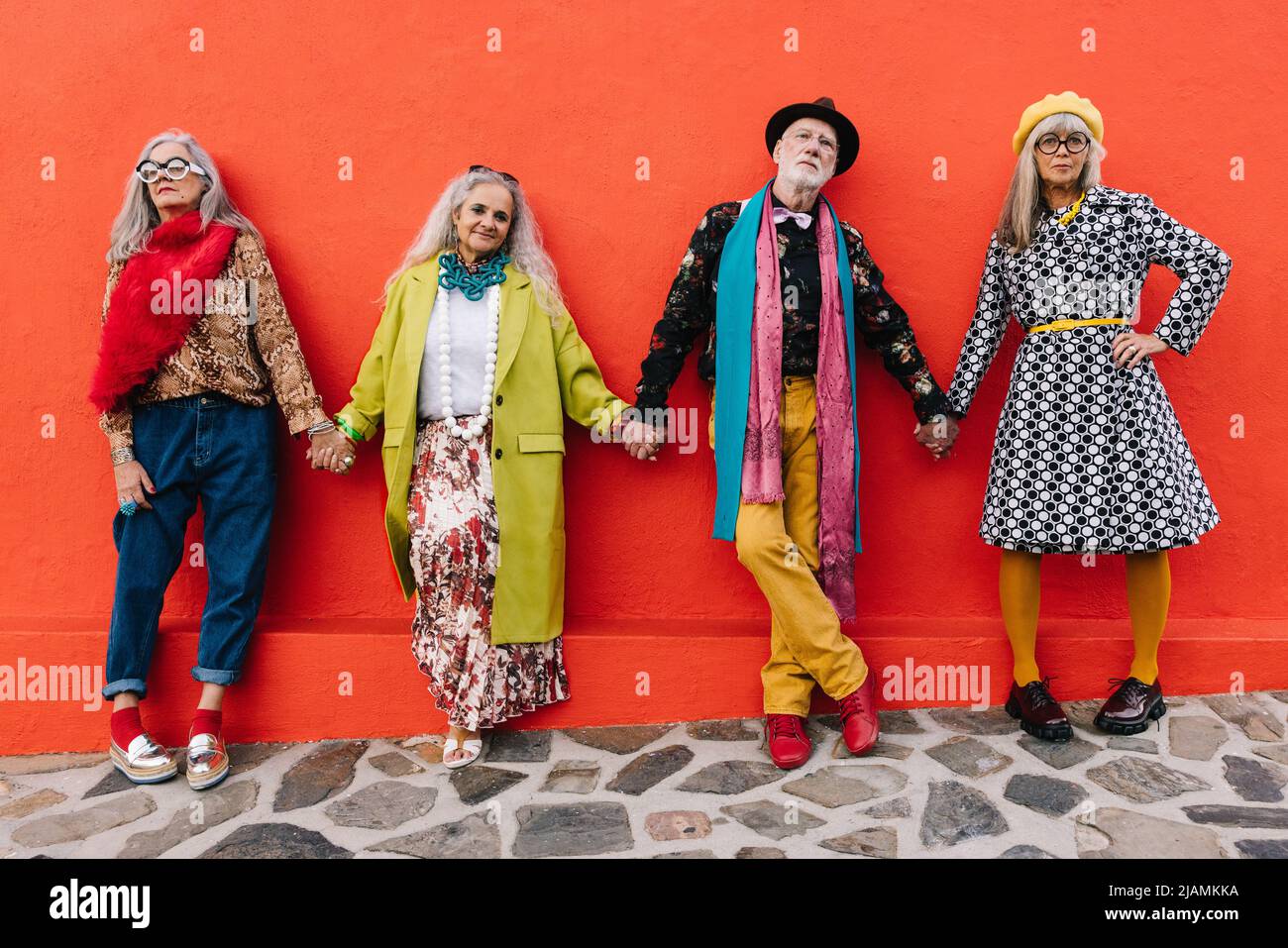 Amici senior che tengono le mani mentre si levano in piedi insieme contro un muro rosso. Quattro persone anziane che esprimono la loro unità e il loro sostegno l'una dell'altra. Gruppo o Foto Stock