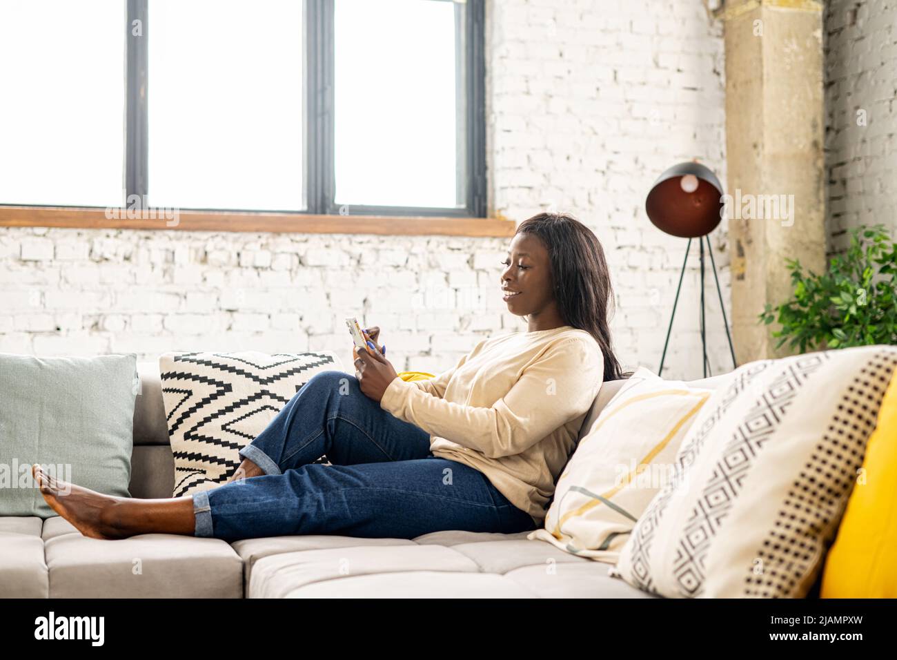 Donna multirazziale che guarda lo smartphone nelle sue mani. Donna che usa il telefono mentre si trova sul divano. Tecnologie e concetto di persone Foto Stock