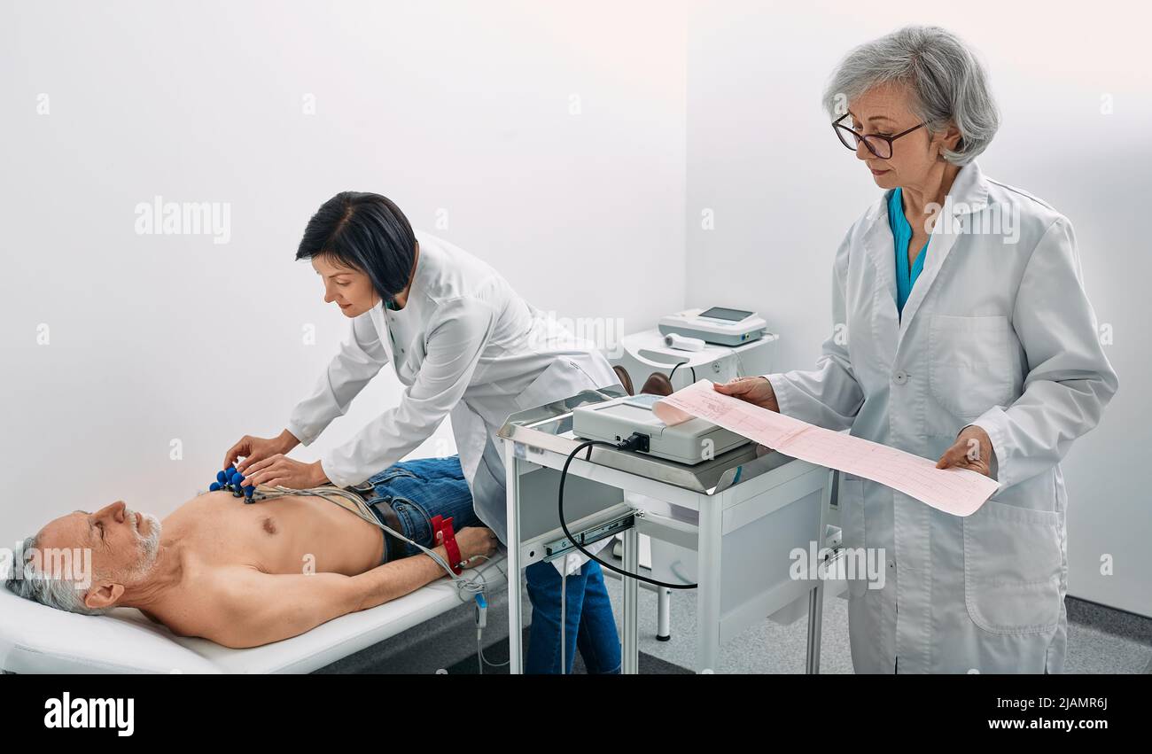 Test del cardiogramma. Il paziente anziano di sesso maschile riceve il monitoraggio della frequenza cardiaca mediante un'apparecchiatura di elettrocardiogramma con due medici cardiologi Foto Stock