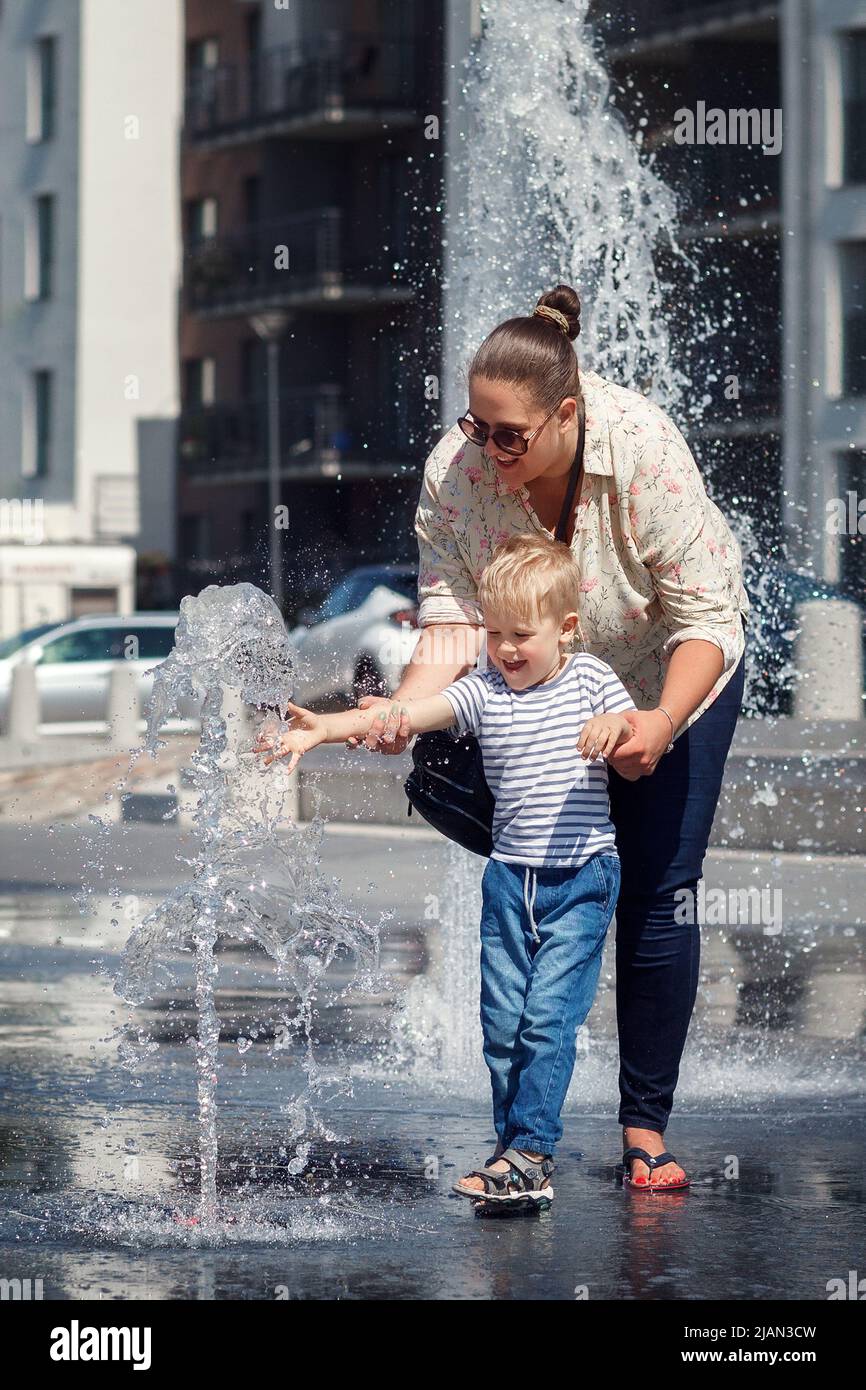 Mamma e suo figlio, nel centro della città, presso una fontana d'acqua. Il bambino ha paura di toccare l'acqua in modo che non si bagna, la madre incoraggia Foto Stock