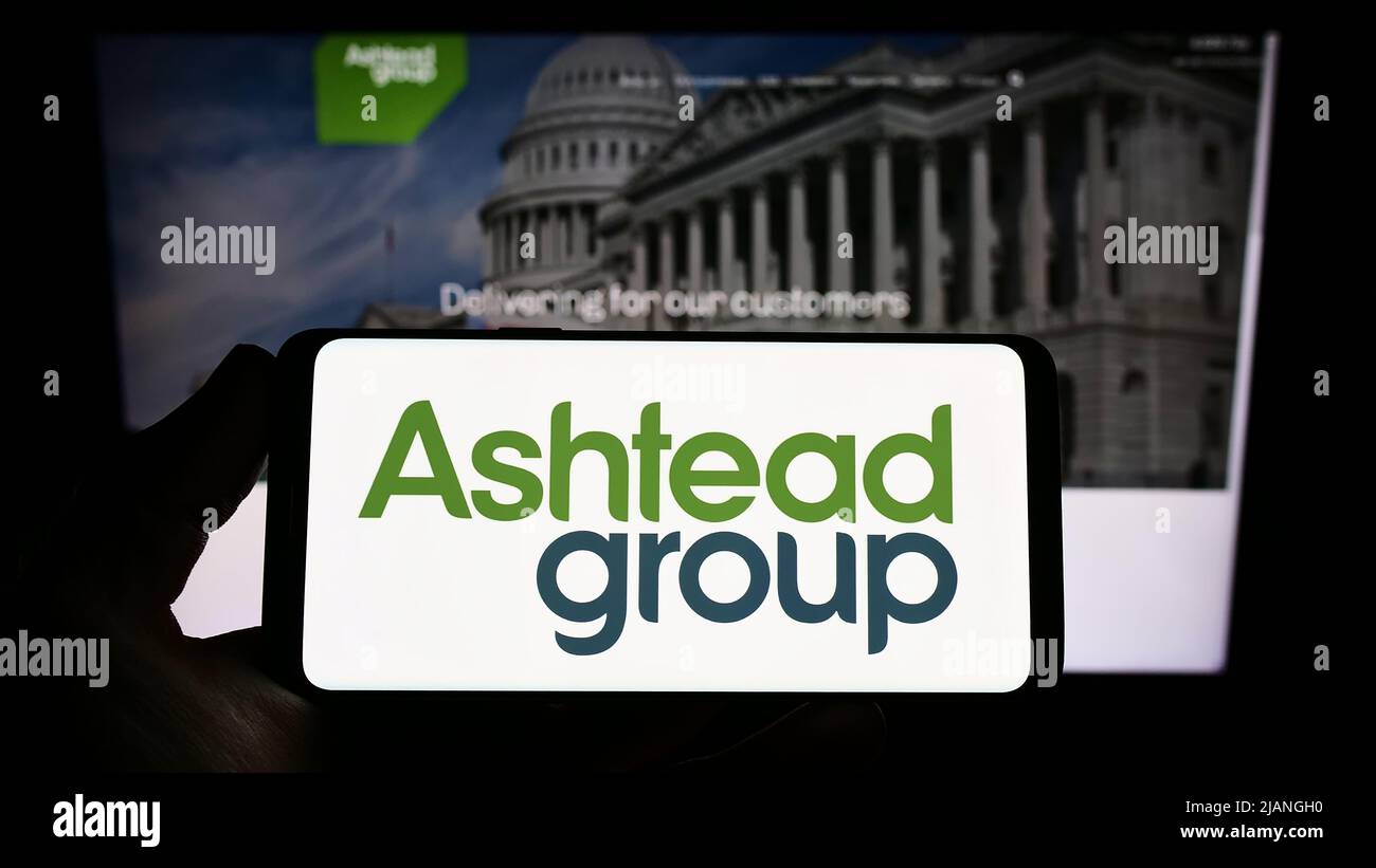 Persona che tiene uno smartphone con il logo della società di noleggio di attrezzature Ashtead Group plc sullo schermo di fronte al sito web. Mettere a fuoco sul display del telefono. Foto Stock