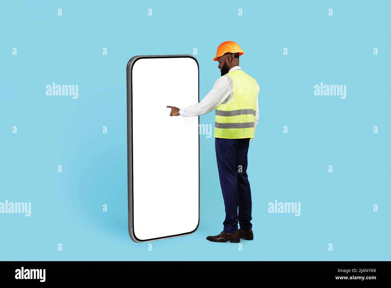 Black Builder in Hard Hat toccare schermo bianco su enorme smartphone vuoto Foto Stock