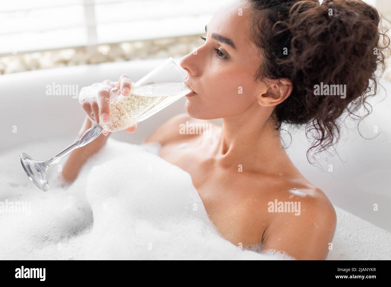 Una signora millenaria sognante beve champagne mentre si rilassa nella vasca a bolle d'aria in un hotel di lusso Foto Stock